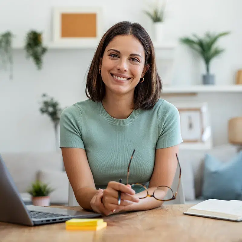 Smiling woman working at home desk with laptop, notebook and glasses Smiling woman working at home desk with laptop, notebook and glasses