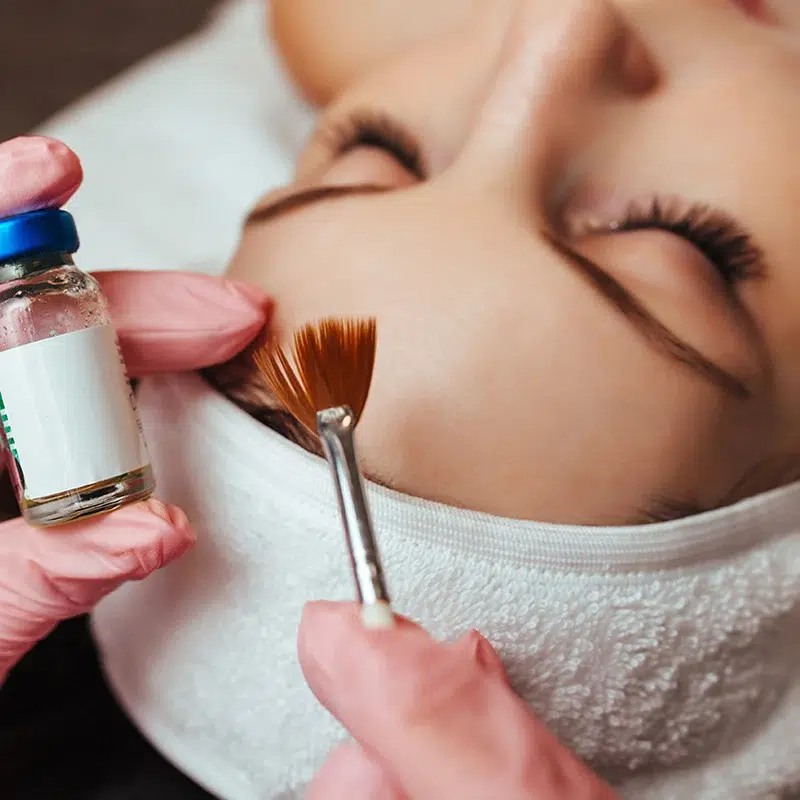 Esthetician in pink gloves applying a biorepeel or serum to a patient's forehead using a brush.